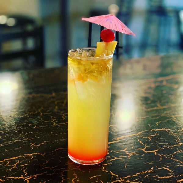 A vibrant yellow and orange cocktail with pineapple, a cherry, and a pink umbrella on a dark marbled table.