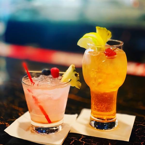 Two colorful cocktails, one pink with berries and pineapple, one orange with lemon and cherry, on a dark table.